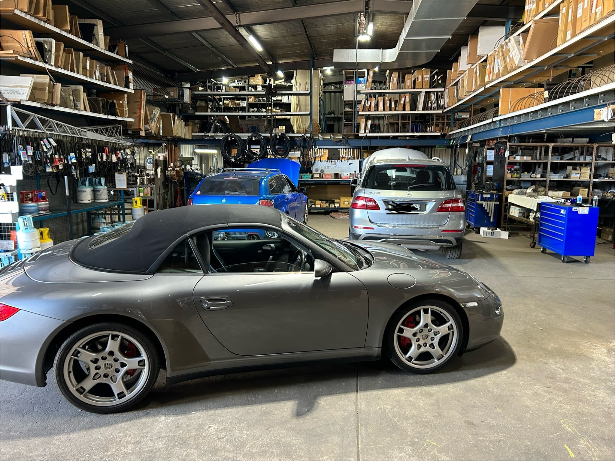 A grey Porsche 911 Cabriolet in the A1 Auto Air parts warehouse, with a Mercedes and stocked shelving behind